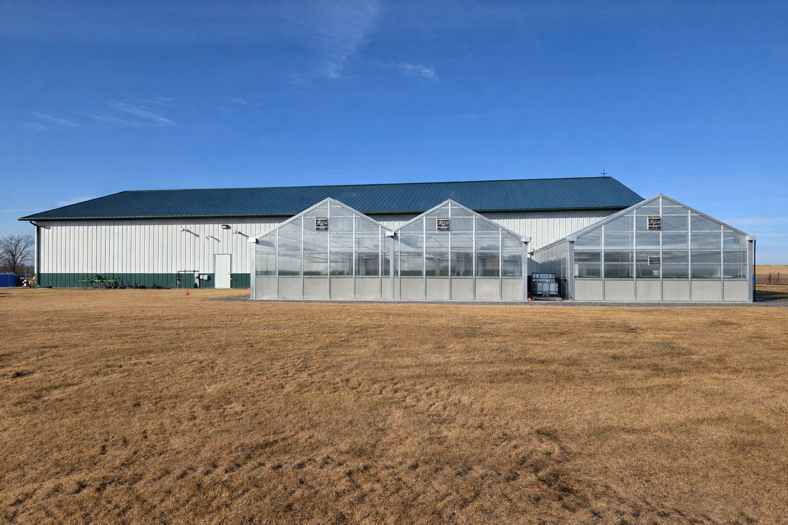 Exterior view of the Legacy Seeds Research & Learning Center in Waupaca, Wisconsin, featuring three newly constructed glass greenhouses attached to the main facility building under a clear blue sky.